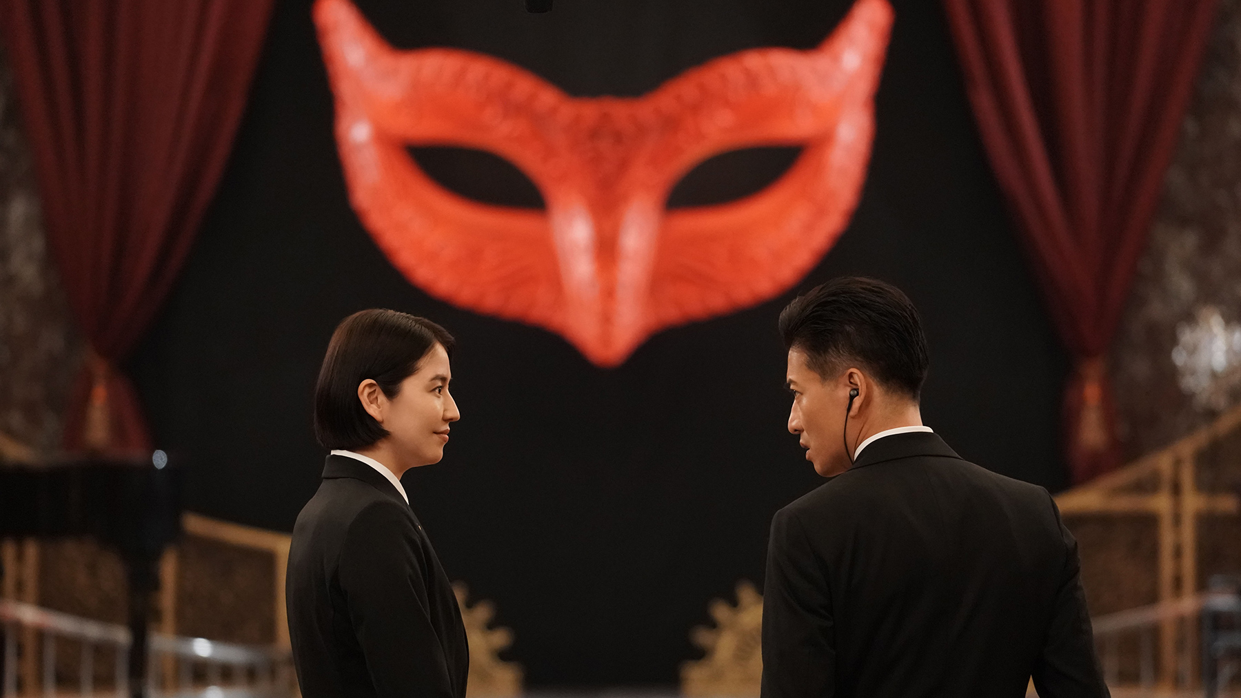 two police detectives talking with large masquerade mask hanging in the background